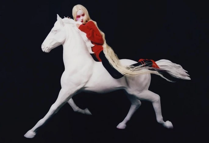Blonde woman in a red top rides a white sculpted horse against a black background in a fashion editorial style photograph.