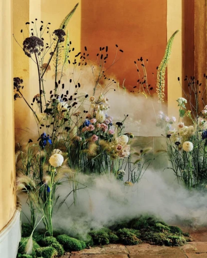 Floral arrangement with white, pink, and blue flowers, green foliage, and moss, creating a soft, misty garden scene inside.