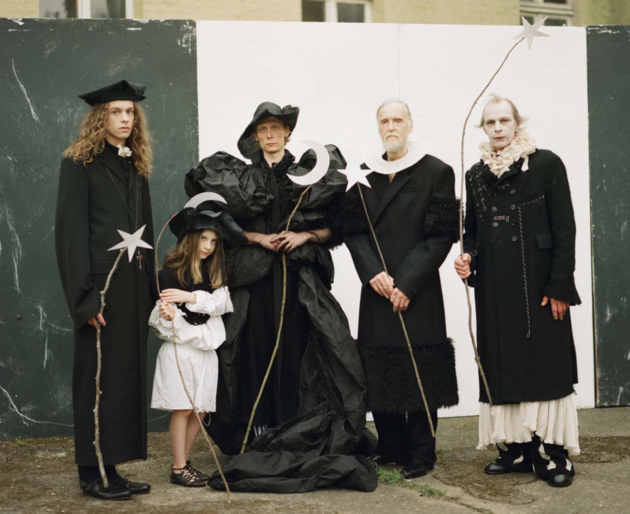 Five people in elaborate black and white costumes stand in front of a weathered black and white wall, holding star and moon props.