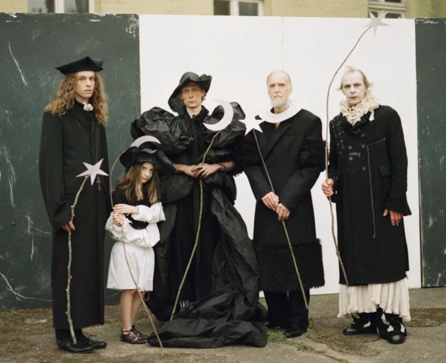 Five people in elaborate black and white costumes stand in front of a weathered black and white wall, holding star and moon props.