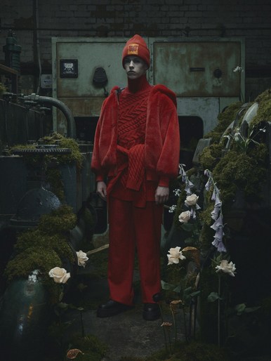 Fashion portrait of a young adult in a red knit outfit with a red beanie, standing among moss-covered industrial objects and white roses.