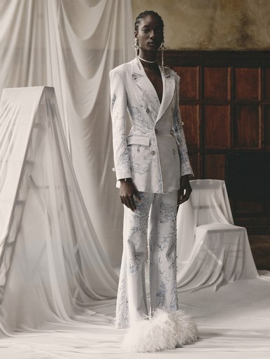 Fashion model in a white, patterned suit with unique furry boots posing in front of draped fabric and antique wood wall.