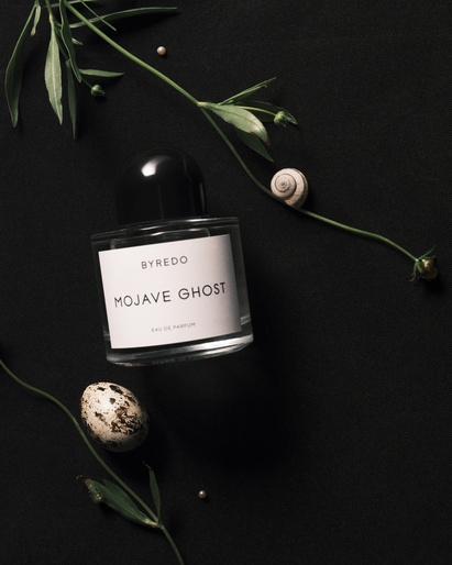 Byredo Mojave Ghost perfume bottle sits on a black surface, adorned with greenery, a snail shell, and a speckled egg.