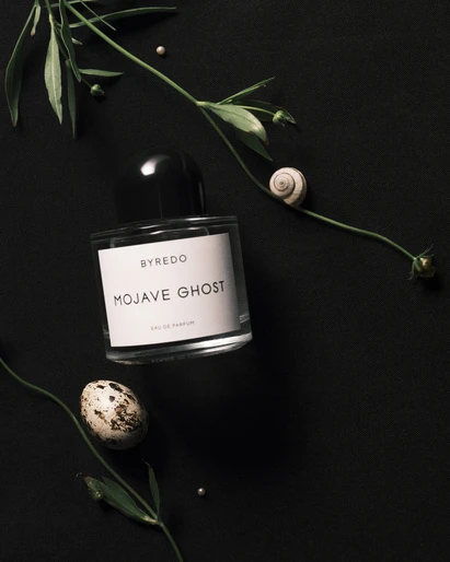 Byredo Mojave Ghost perfume bottle sits on a black surface, adorned with greenery, a snail shell, and a speckled egg.