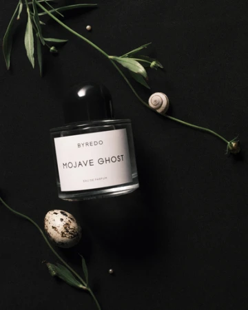 Byredo Mojave Ghost perfume bottle sits on a black surface, adorned with greenery, a snail shell, and a speckled egg.