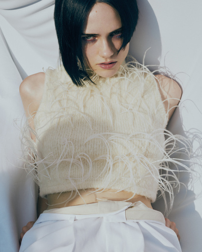 Model with dark hair wearing a feather-trimmed white top and light trousers against a bright white background.