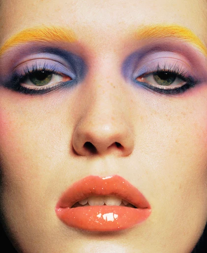 Close-up beauty portrait of a woman with vibrant yellow eyebrows, purple eyeshadow, and glossy coral lips by Olivia Giles