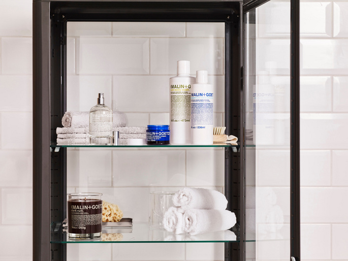 Bathroom cabinet showcases Malin+Goetz products: candles, cleansers, and towels against white tiled wall. Focus on beauty and organization.