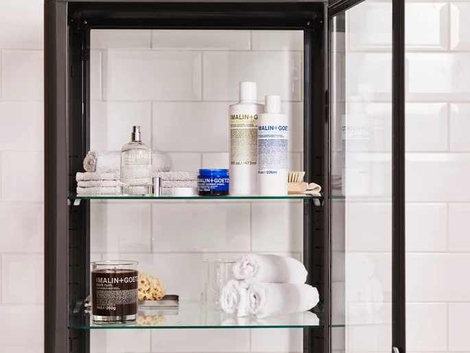 Bathroom cabinet showcases Malin+Goetz products: candles, cleansers, and towels against white tiled wall. Focus on beauty and organization.