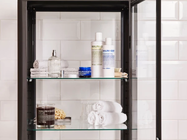 Bathroom cabinet showcases Malin+Goetz products: candles, cleansers, and towels against white tiled wall. Focus on beauty and organization.
