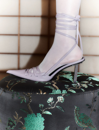 Elegant lavender heeled shoe on a patterned ottoman, paired with sheer socks, stands out against a neutral grid background.