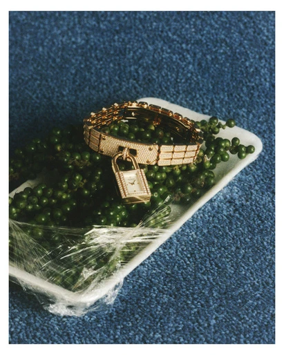 High-angle studio shot: gold watch & bracelet on a white tray filled with green peppercorns, covered in plastic wrap, against a blue carpet.