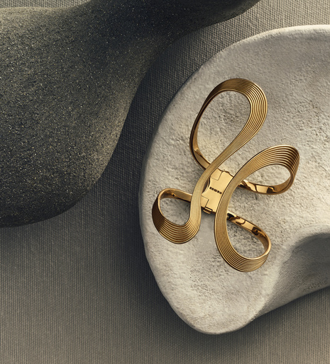 Close-up of an elegant gold bracelet with abstract loops resting on a textured grey surface, complemented by grey sculptural forms.