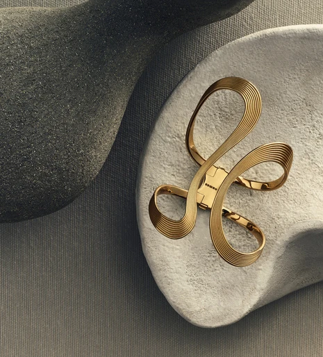 Close-up of an elegant gold bracelet with abstract loops resting on a textured grey surface, complemented by grey sculptural forms.