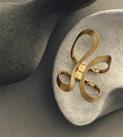 Close-up of an elegant gold bracelet with abstract loops resting on a textured grey surface, complemented by grey sculptural forms.