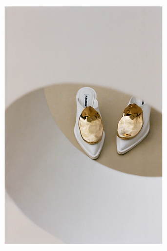 Elevated studio shot of a pair of white heeled mules with large golden decorative elements, set against a neutral beige surface.