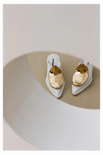 Elevated studio shot of a pair of white heeled mules with large golden decorative elements, set against a neutral beige surface.