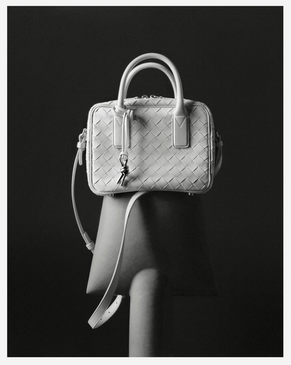 White woven leather handbag with double top handles and shoulder strap on a chair against a dark grey background in a studio shot.