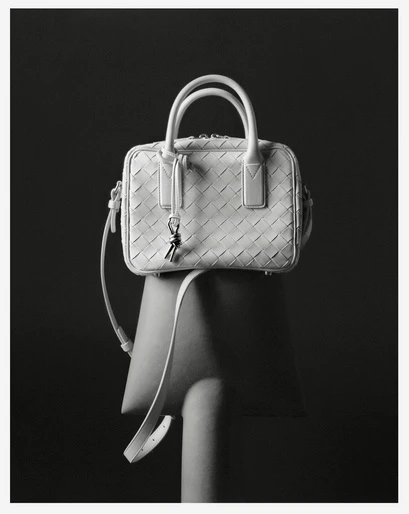 White woven leather handbag with double top handles and shoulder strap on a chair against a dark grey background in a studio shot.