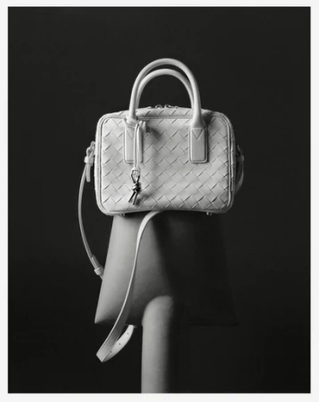 White woven leather handbag with double top handles and shoulder strap on a chair against a dark grey background in a studio shot.