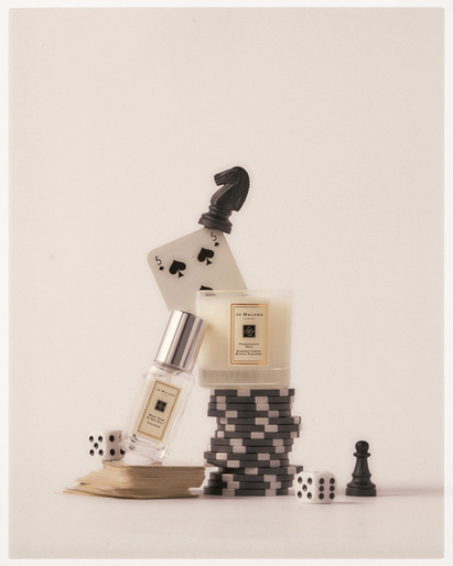 Still life shot of perfume bottle, candle, dice, poker chips, playing card, and chess pieces against a light-beige seamless backdrop.
