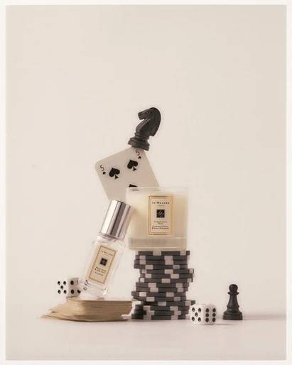 Still life shot of perfume bottle, candle, dice, poker chips, playing card, and chess pieces against a light-beige seamless backdrop.