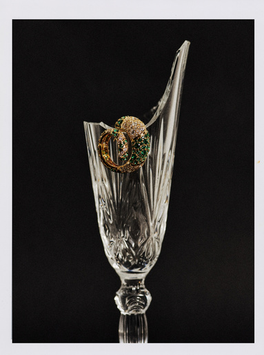 Crystal glass with a broken rim showcases ornate gold hoop earrings with green and white stones against a stark black background.