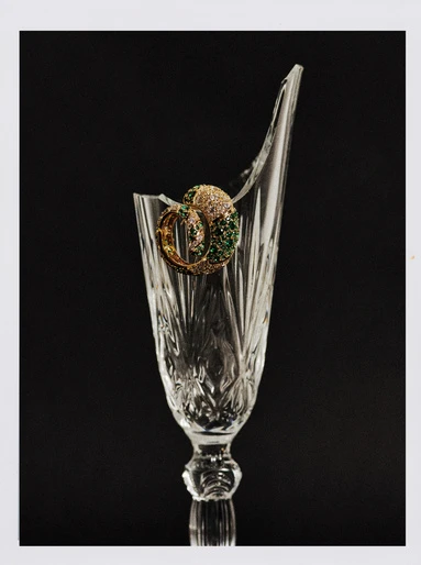 Crystal glass with a broken rim showcases ornate gold hoop earrings with green and white stones against a stark black background.