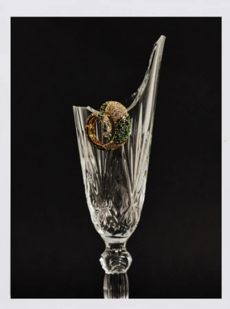 Crystal glass with a broken rim showcases ornate gold hoop earrings with green and white stones against a stark black background.