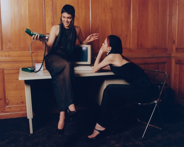 Two women in elegant black dresses pose near a vintage computer and green phone. One woman sits on the desk holding the phone.