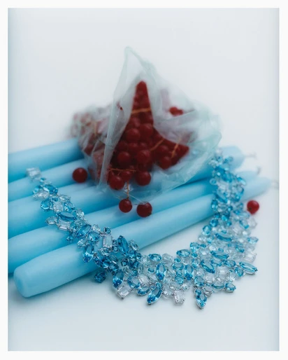 Still life featuring a crystal necklace draped over light blue candles, with red currants in a plastic bag in a bright, soft-lit setting.