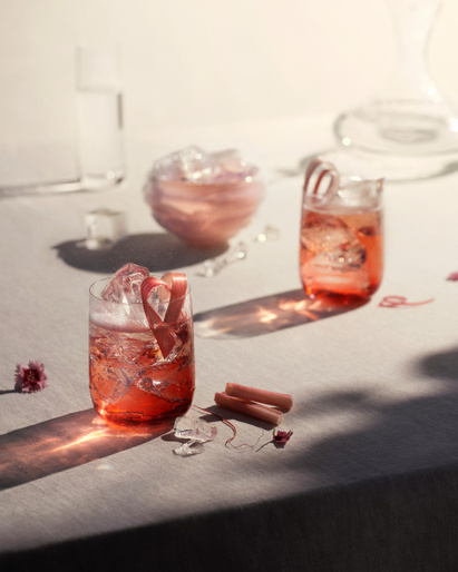 Two glasses of red cocktails with ice and rhubarb garnishes on a table with glass pitchers and flowers