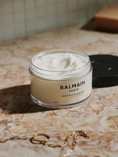 Balmain Paris hair mask product shot on a marble surface with a gold chain necklace and warm, diffused lighting.