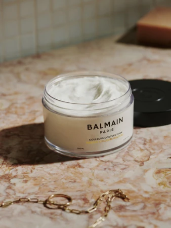 Balmain Paris hair mask product shot on a marble surface with a gold chain necklace and warm, diffused lighting.