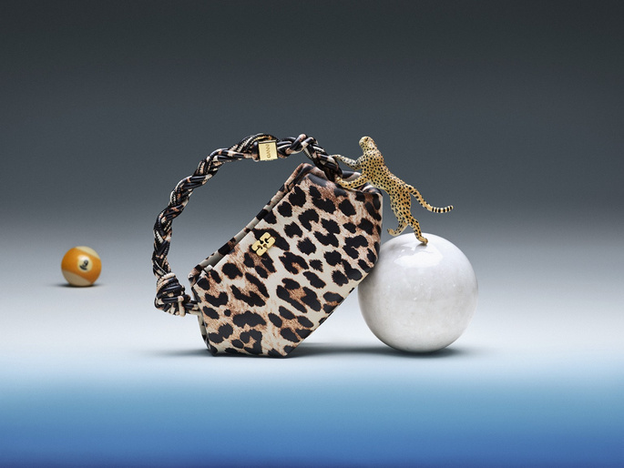 Leopard print handbag with a braided handle next to a toy cheetah on a sphere with a billiard ball in the neutral gradient background.