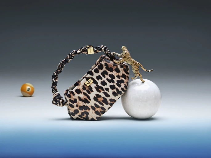 Leopard print handbag with a braided handle next to a toy cheetah on a sphere with a billiard ball in the neutral gradient background.