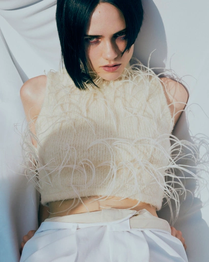 A woman with dark hair wearing a cream feather-trimmed top and white trousers against a white background by Lucy Blofeld