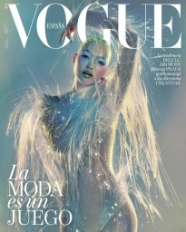 Model with shaved head wearing a shimmering sheer embellished bodysuit with ethereal wispy fiber attachments against a muted green gradient background.