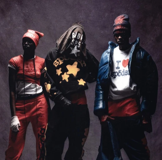 Three people wearing streetwear fashion, including hooded jackets and hats, pose in front of a dark gray background in a studio shot.