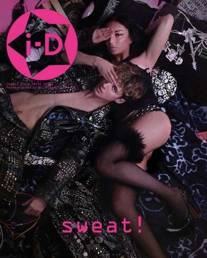 Fashion editorial shot: Two models pose together on a bed. One model covers their face, wearing a studded jacket and black stockings.