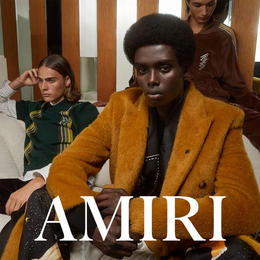 Three fashion models showcase AMIRI clothing in a casual indoor setting, featuring a golden jacket, patterned shirt, and stylish bottoms.