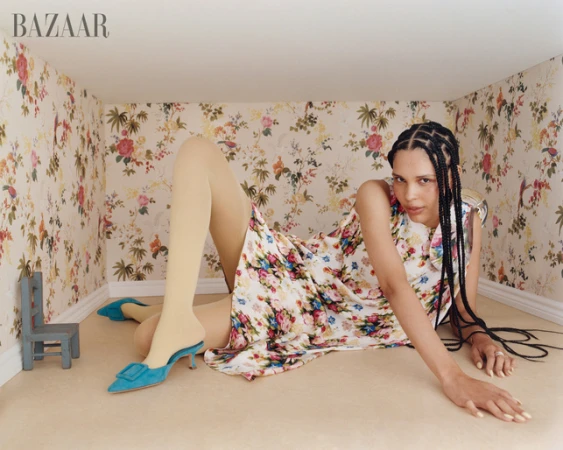 Fashion editorial shot of a woman in a floral dress and tights laying in a room with floral wallpaper and a small blue chair.