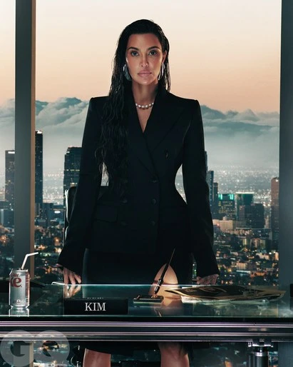 Portrait of Kim Kardashian in a black business suit at a desk in front of a high-rise window with a city view at dusk.