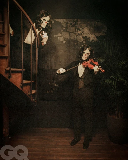 Two people wearing skeleton makeup and formal wear, one playing the violin, posing indoors in dim lighting with a spiral staircase.