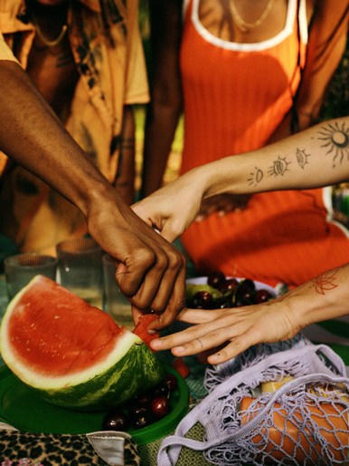Three people gathered around a picnic table, with a slice of watermelon, cherries, and a reusable shopping bag filled with fruit.