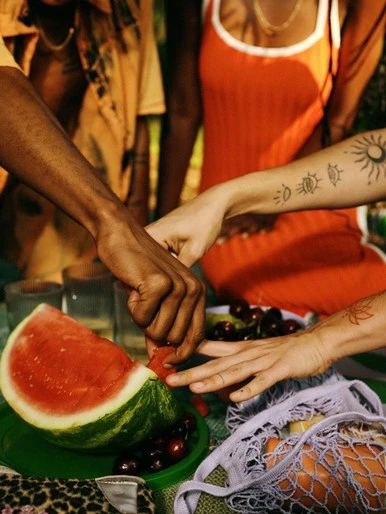 Three people gathered around a picnic table, with a slice of watermelon, cherries, and a reusable shopping bag filled with fruit.