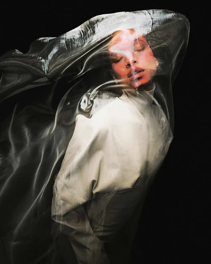 Woman with eyes closed stands draped in a translucent, shimmering fabric, posing against a dark background in a fashion-style portrait.