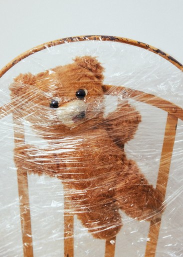 A brown teddy bear is tied to a wooden chair with clear plastic wrap in a well-lit indoor setting. The plastic wrap covers the bear.