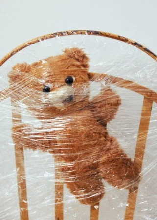 A brown teddy bear is tied to a wooden chair with clear plastic wrap in a well-lit indoor setting. The plastic wrap covers the bear.