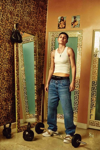 Person in tank top and jeans poses in room with weights and mirrors. Interior has leopard-print wallpaper and Greek-key trim.